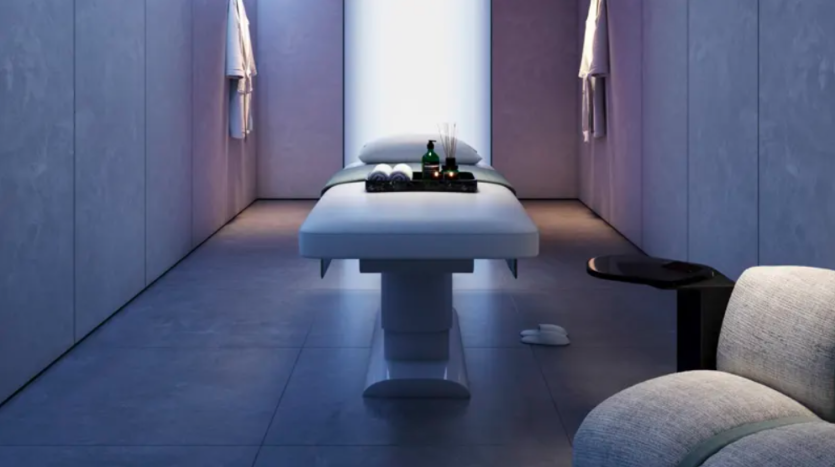 Une salle de spa minimaliste et faiblement éclairée comprend une table de massage avec des serviettes, des huiles essentielles et des articles d'aromathérapie bien rangés. Les murs sont lisses et gris et une chaise moelleuse se trouve dans le coin, soulignant une atmosphère tranquille.