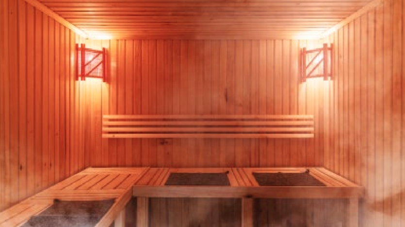 Un sauna en bois au sein du W Residences Dubai Harbour présente des lumières chaudes et lumineuses sur les murs et de la vapeur s'élevant du sol. Des bancs en bois bordent les murs sous un plafond à lattes en bois. L'ambiance est chaleureuse et accueillante, parfaite pour se détendre dans le luxe.