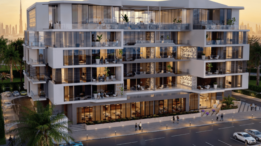 Un bâtiment moderne de plusieurs étages dans les jardins d'Ayana, doté de grandes fenêtres en verre et de balcons, est éclairé par un éclairage chaleureux pendant la soirée. Les gens marchent sur le trottoir et les voitures sont garées dans la rue. Les toits de la ville sont visibles en arrière-plan et des palmiers bordent la zone.