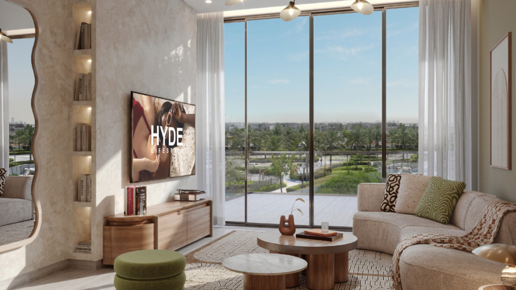 Salon moderne des Hyde Residences avec de grandes baies vitrées offrant une vue sur un paysage verdoyant. La pièce est décorée d'un canapé beige, d'un pouf vert, de tables basses rondes et d'une télévision murale affichant « HYDE ». Un éclairage doux et des tons neutres créent une atmosphère chaleureuse.