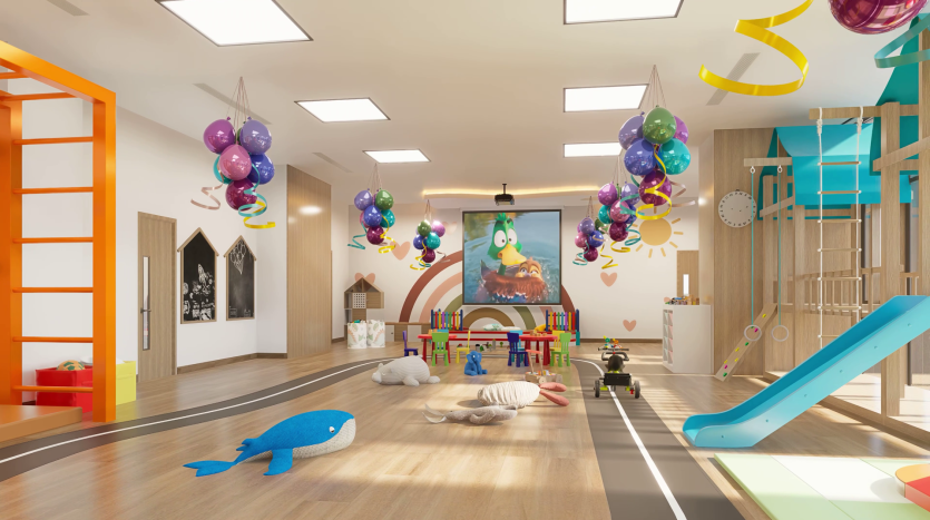 Une salle de jeux bien éclairée avec des décorations colorées, des ballons et des banderoles suspendues au plafond. Diverses aires de jeux comprennent des toboggans, des portiques d'escalade, des véhicules jouets et une grande table avec des chaises. Des peluches comme une baleine bleue et d'autres animaux en peluche sont posées au sol.