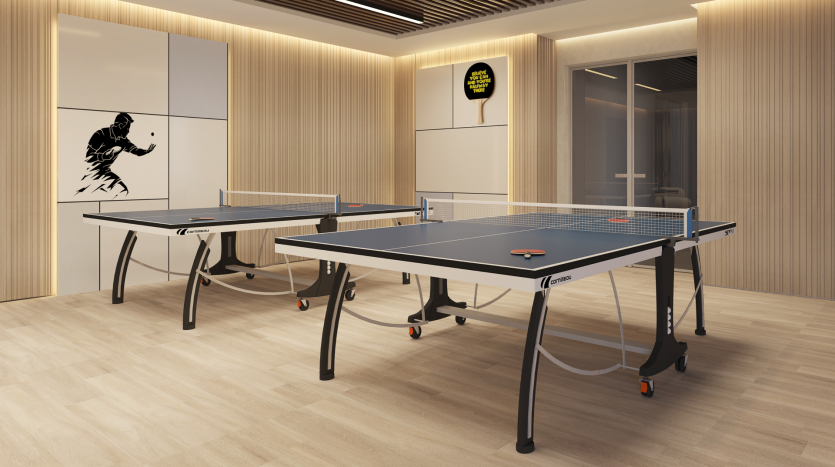 Une salle moderne avec deux tables de ping-pong installées pour jouer. Les murs sont décorés de boiseries, d'une fresque murale représentant un joueur de tennis de table et d'un panneau indiquant "Game On". La pièce dispose d'un éclairage suffisant et d'une porte vitrée en arrière-plan.