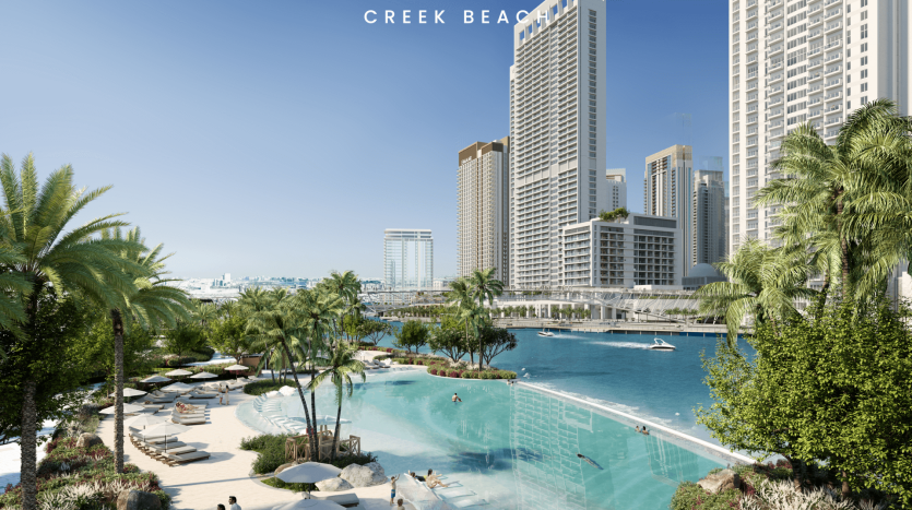 Une vue pittoresque sur Creek Beach avec une plage de sable sereine avec des parasols et des chaises longues, des palmiers et un lagon bleu calme. Des bâtiments hauts et modernes se profilent à l’arrière-plan sous un ciel bleu clair, tandis qu’un yacht est amarré à proximité.