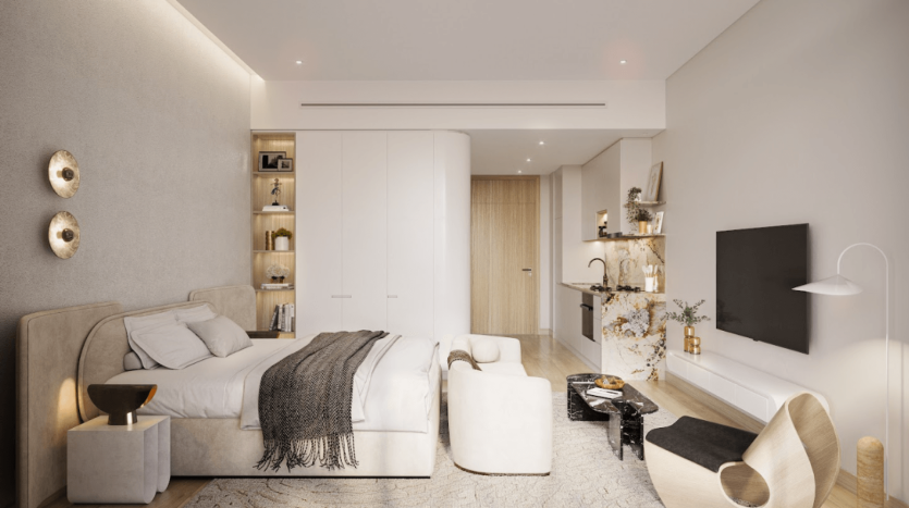 Une chambre moderne du One Residences comprend un lit moelleux aux tons neutres, un jeté gris et des tables de chevet. Les étagères et armoires intégrées offrent un grand espace de rangement. Un coin salon confortable avec une table, des chaises et une télévision se trouve près de la porte. Un éclairage doux et des éléments de décoration créent une ambiance chaleureuse et accueillante.