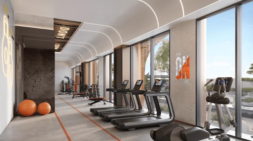 Une salle de sport moderne à One Residences dispose de grandes fenêtres et d'une lumière naturelle abondante. L'équipement comprend des tapis roulants, des vélos elliptiques, des appareils de musculation et des ballons d'exercice. Les murs sont ornés de décalcomanies motivantes, avec une vue sur les arbres et un ciel bleu clair à l'extérieur.