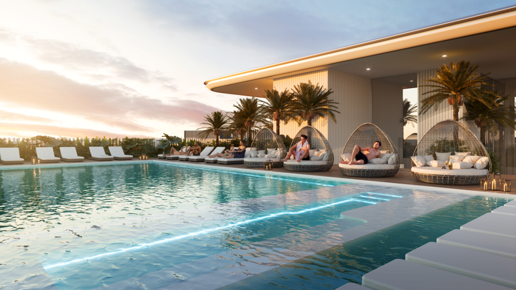 Une piscine à débordement sur le toit avec une eau cristalline et des chaises longues. Les gens se détendent sur des chaises gigognes à l’ombre des palmiers. La scène se déroule au coucher du soleil, projetant une lueur chaleureuse sur le bâtiment moderne Cove Edition One en arrière-plan.