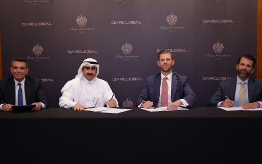 Quatre hommes en costume sont assis à une table avec une nappe sombre, devant un fond affichant les logos "DarGlobal" et "Trump International". Trois des hommes tiennent un stylo comme s'ils signaient des documents, tandis que l'homme à l'extrême gauche sourit à la caméra, contre le projet de Trump Tower Jeddah.