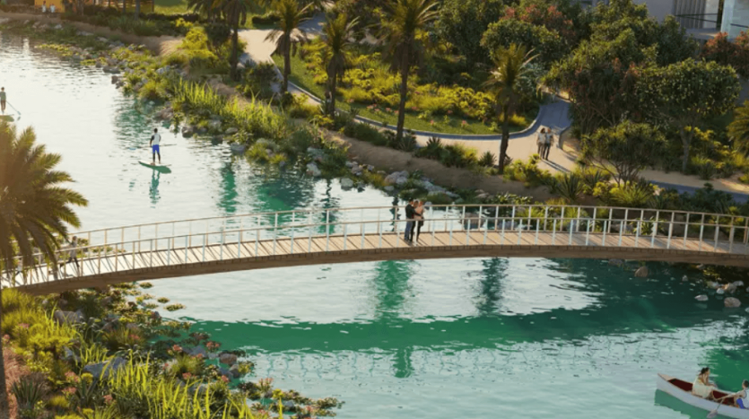 Un parc pittoresque comprend un canal aux eaux bleues claires et une végétation luxuriante le long des berges. Des gens marchent sur un pont, une personne fait du paddleboard sur l’eau violette et une autre rame sur un petit bateau. Les arbres et les sentiers ajoutent à l’ambiance extérieure vibrante.