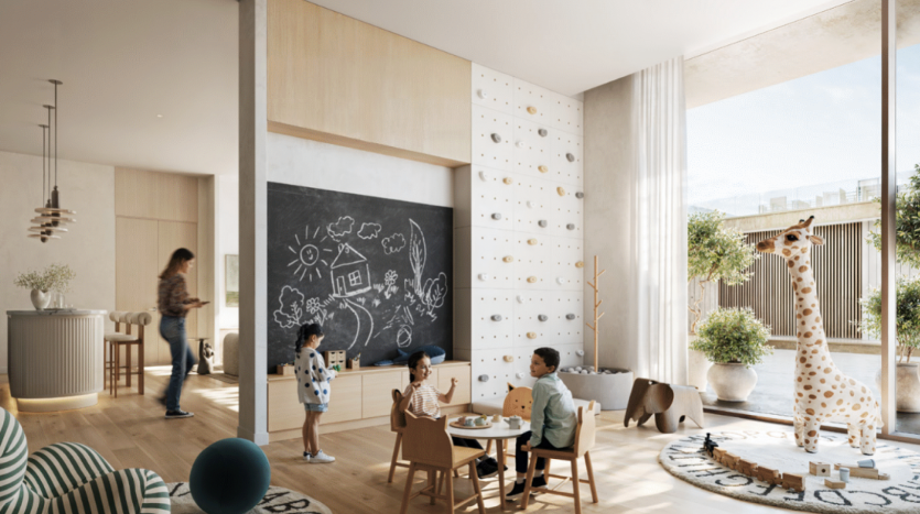 La salle de jeux pour enfants lumineuse et moderne de Address Residences comprend un mur en tableau noir couvert de dessins, un mur d'escalade et un tapis rond avec une grande girafe en peluche. Deux enfants sont assis à une petite table avec une femme en arrière-plan et une cuisine visible à gauche.