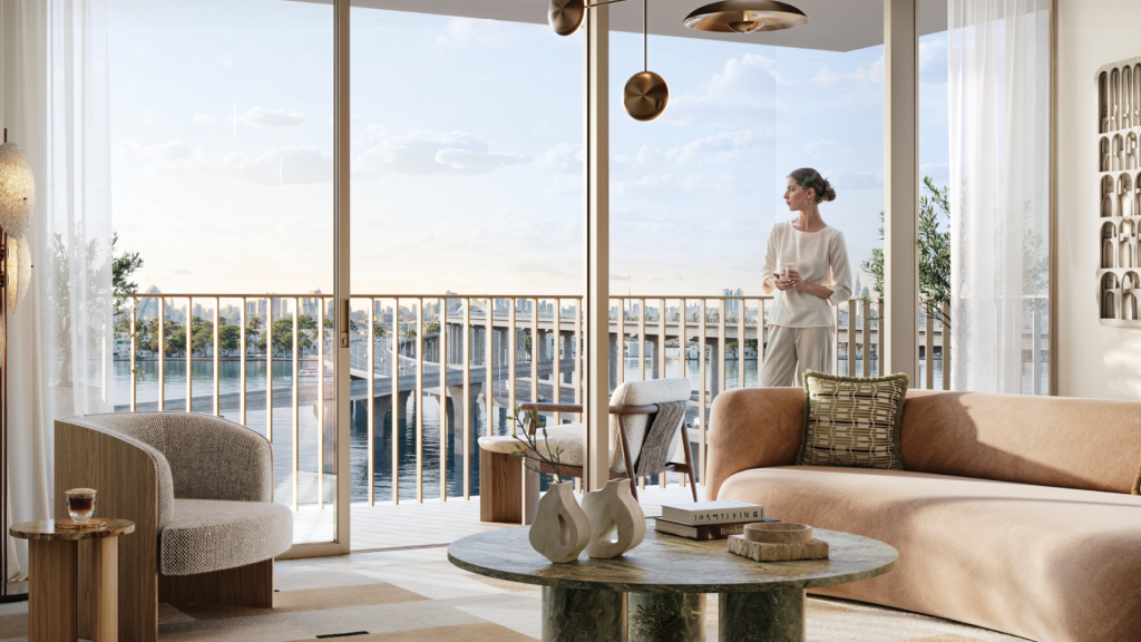 Une personne se tient sur un balcon de l'Address Residences, surplombant un front de mer serein. À l'intérieur, le salon dispose d'un mobilier élégant comme un canapé beige, une table basse en marbre vert avec décoration, une chaise rembourrée et des luminaires élégants. De grandes fenêtres laissent entrer beaucoup de lumière naturelle.