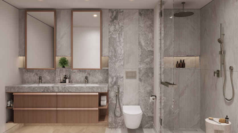 A modern bathroom at Belgrove Residences features light-gray marble walls and fixtures, a double sink with wooden drawers, a wall-mounted toilet, a glass-enclosed shower with rainfall showerhead and built-in shelves containing toiletries and decorative items.