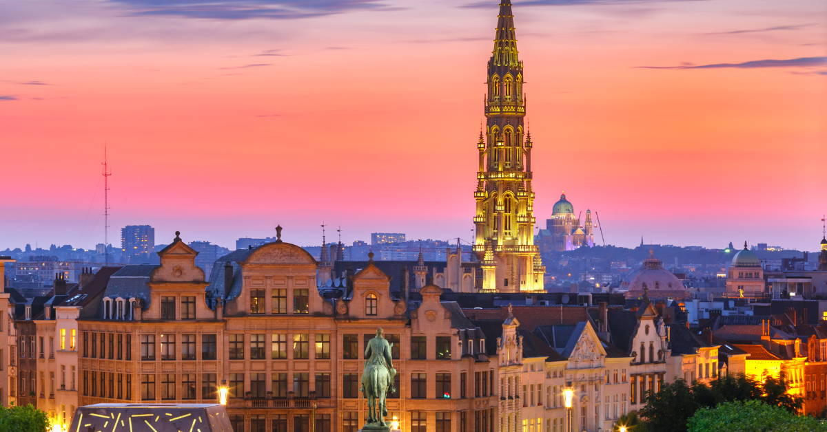 Vue de Bruxelles au coucher du soleil, mettant en valeur l'architecture historique et l'imposante flèche illuminée de l'Hôtel de Ville. Le ciel passe du rose à l'orange, projetant une lueur chaleureuse sur l'horizon de la ville avec une statue visible au premier plan, ressemblant à un salon en plein air d'une beauté intemporelle.