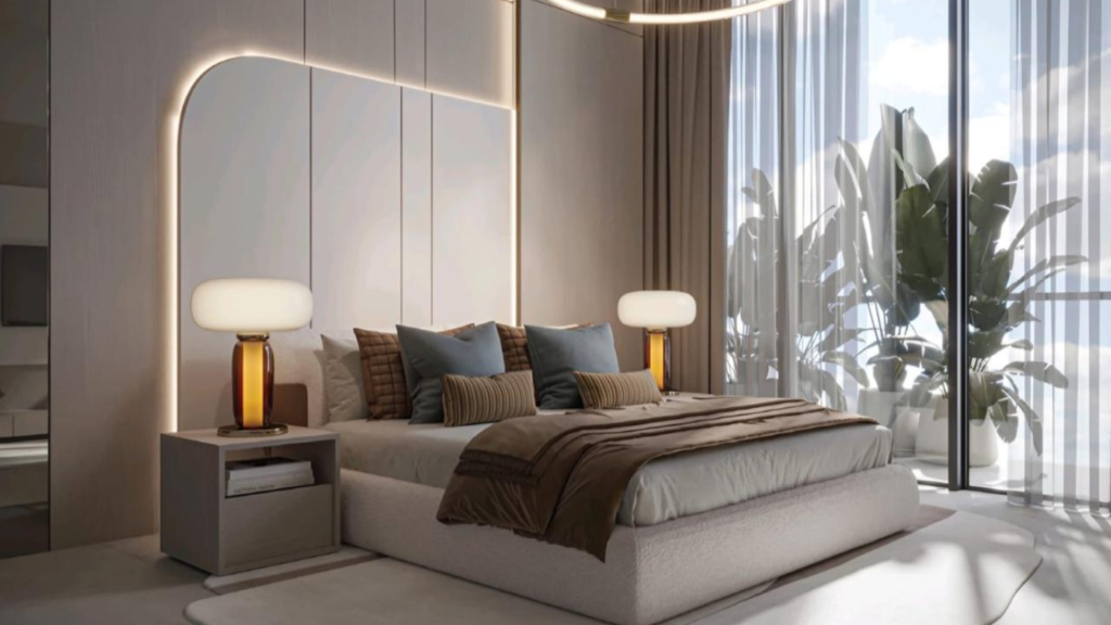 Modern room with large windows and sheer curtains letting in lots of natural light. The room features a soft bed with brown, beige and blue pillows from the Legado Prescott collection, two bedside tables with amber glass lamps and large potted plants near the windows. A warm, welcoming atmosphere.
