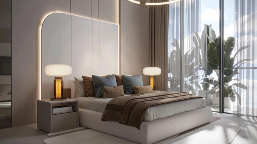 Modern room with large windows and sheer curtains letting in lots of natural light. The room features a soft bed with brown, beige and blue pillows from the Legado Prescott collection, two bedside tables with amber glass lamps and large potted plants near the windows. A warm, welcoming atmosphere.
