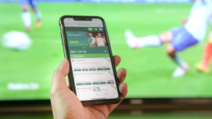 Une main tient un smartphone affichant une application de paris sportifs inspirée du casino, avec en arrière-plan un match de football en direct sur un téléviseur.