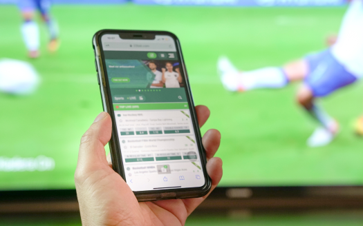 Une main tient un smartphone affichant une application de paris sportifs inspirée du casino, avec en arrière-plan un match de football en direct sur un téléviseur.