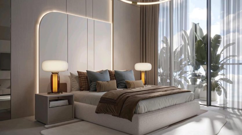 A modern bedroom with a large Legado Prescott bed, flanked by two bedside tables, each supporting a lamp with an amber-colored cylindrical base and white shade. The room features an elegant, light-colored headboard, picture windows with sheer curtains and potted plants.