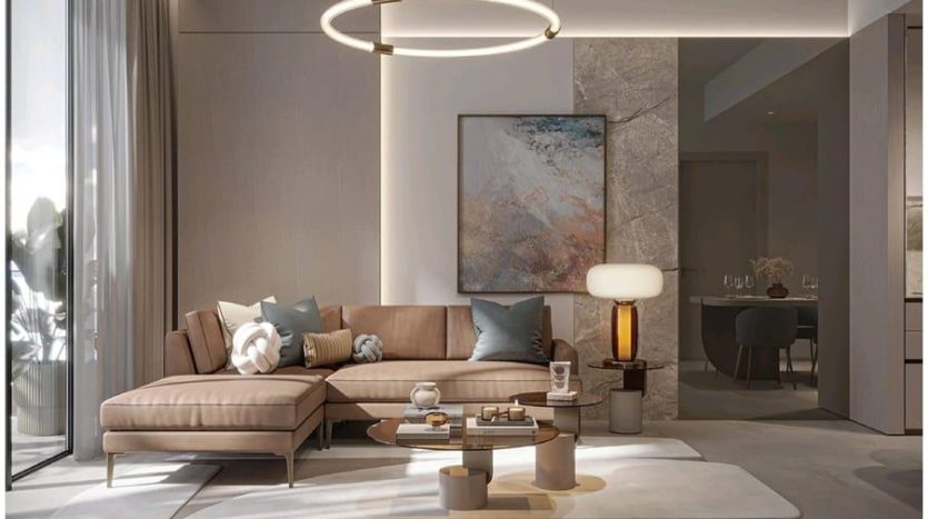 A modern living room in neutral tones features a beige L-shaped Legado Prescott sofa adorned with cushions, a round coffee table and a large abstract painting on a stone accent wall. Lighting includes a circular chandelier and a contemporary table lamp.