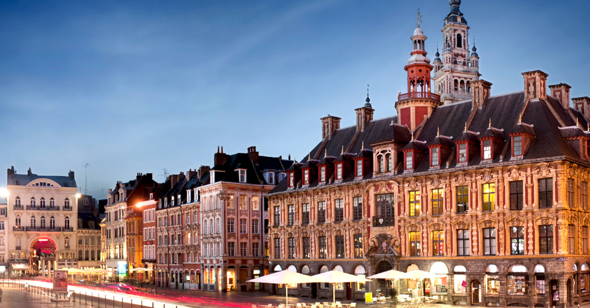 Une place animée au crépuscule présente les grands bâtiments historiques du Salon de Lille aux façades complexes et aux fenêtres illuminées. Le ciel est clair et un éclairage doux met en valeur les détails architecturaux. Quelques traînées lumineuses floues provenant de véhicules en mouvement sont visibles au premier plan.