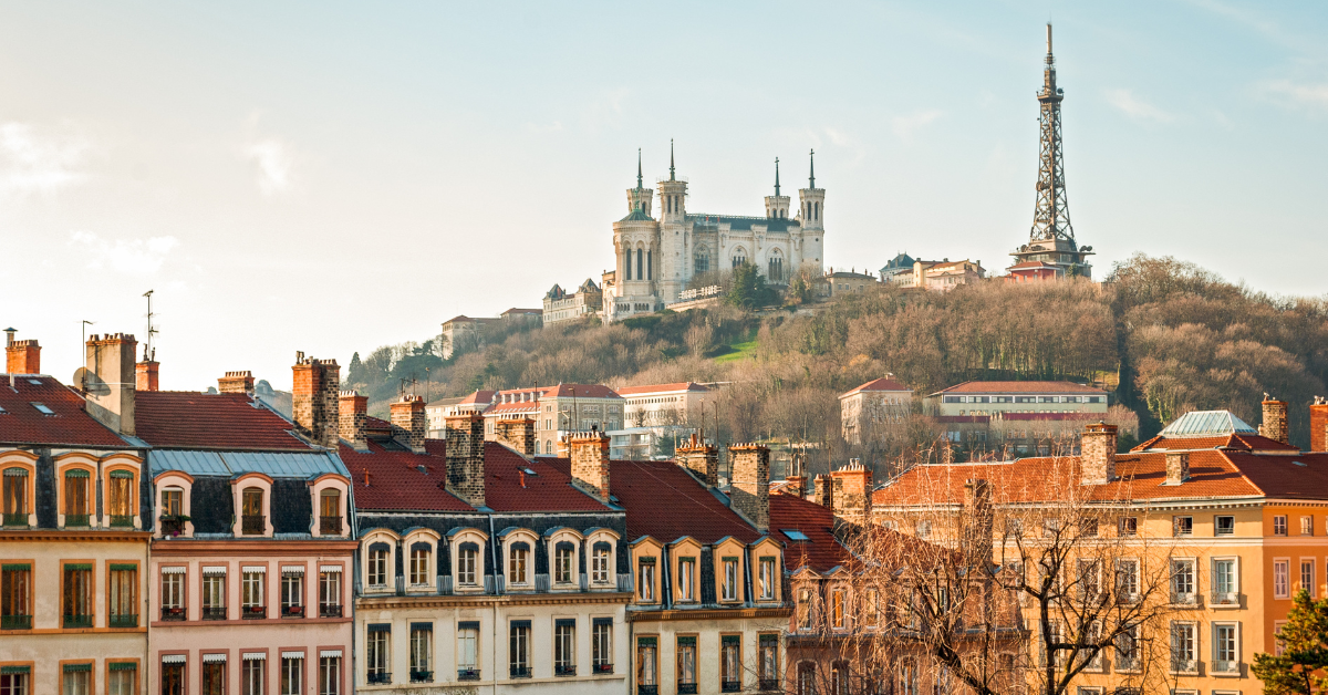 Vue panoramique de Lyon, en France, avec au premier plan un panorama de bâtiments historiques colorés. Au sommet d'une colline se dresse la basilique Notre-Dame de Fourvière avec ses quatre tours, à côté de la Tour Métallique de Fourvière. Le ciel clair ajoute à cette scène pittoresque de brouillon automobile.