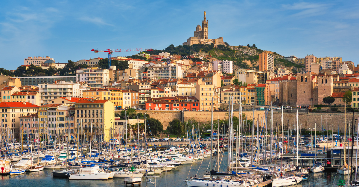 Vue panoramique d'une marina remplie de bateaux amarrés dans un port, avec un paysage urbain de bâtiments colorés et d'architecture côtière en arrière-plan. Une colline avec une église proéminente au sommet de la colline domine l'horizon de Marseille sous un ciel bleu clair.