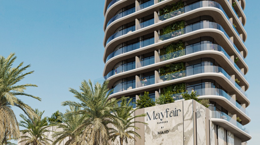 Un immeuble moderne de grande hauteur nommé « Mayfair Gardens by MAJD » avec des balcons incurvés et de vastes fenêtres en verre. La structure est entourée de palmiers luxuriants et des plantes sont visibles sur plusieurs balcons. Le ciel est clair et bleu.