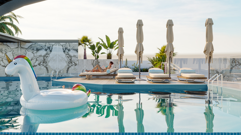 Une piscine sur le toit avec une grande licorne blanche flottant dans l'eau. Des chaises longues avec des parasols beiges pliés sont alignées au bord de la piscine, et une personne se détend sur l'une des chaises longues. L'arrière-plan présente une végétation luxuriante et un mur en marbre.