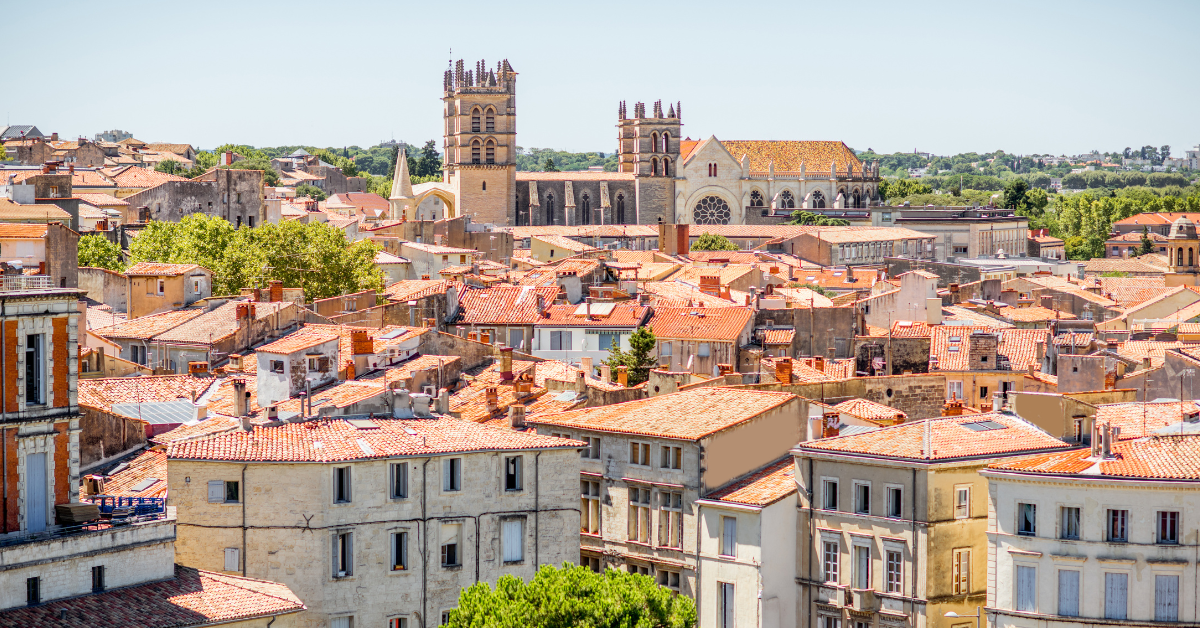 Vue panoramique d'une ville européenne historique aux toits de tuiles rouges et aux bâtiments beiges. En arrière-plan se dresse une grande cathédrale de style gothique avec deux clochers. Des arbres et de la verdure sont visibles entre les bâtiments par une journée ensoleillée, rappelant le salon immobilier de Dubaï à Montpellier.