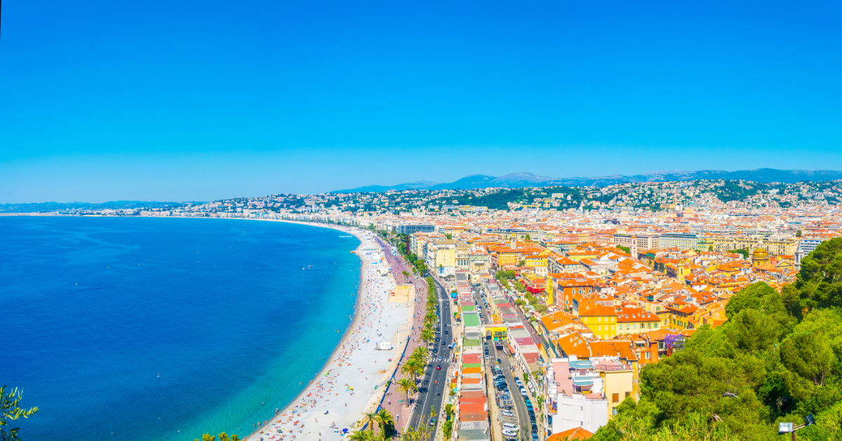 Un paysage urbain côtier à Nice avec une longue plage de sable aux eaux bleues claires qui s'étendent jusqu'à l'horizon. La beauté de la plage est complétée par les bâtiments et les routes qui bordent son bord, tandis que des collines verdoyantes et d'autres structures remplissent l'arrière-plan sous un ciel bleu éclatant.