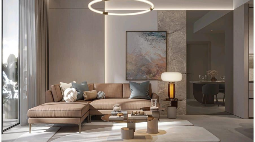 A modern living room with a beige corner sofa adorned with gray and white cushions, a round coffee table, a large abstract painting on the wall and a circular hanging lamp. The Legado Prescott-themed space features a large window with sheer curtains and an adjacent dining room visible in the background.