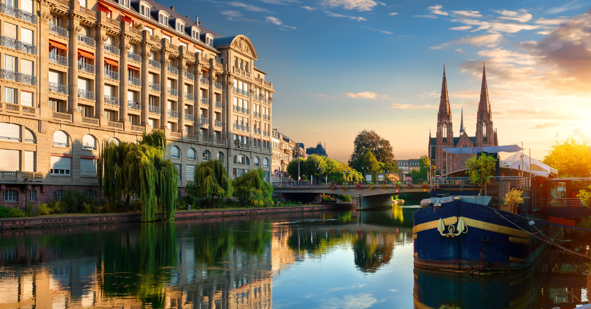 Une scène sereine de canal à Strasbourg avec des reflets de bâtiments historiques et de flèches dans l'eau. À gauche, une architecture grandiose avec des balcons et de grandes fenêtres borde le canal, rappelant un salon élégant, tandis qu'à droite, un bateau est amarré avec des flèches et un pont en arrière-plan sous un ciel clair et coloré.