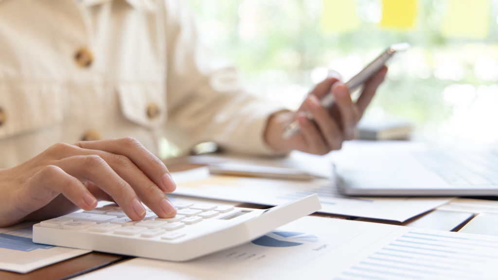 Una persona in camicia chiara utilizza una calcolatrice bianca su una scrivania ingombra di documenti e un computer portatile, tenendo in mano uno smartphone. La scena fa pensare a un lavoro d'ufficio o a calcoli finanziari, forse legati alla vendita di un appartamento a Dubai.