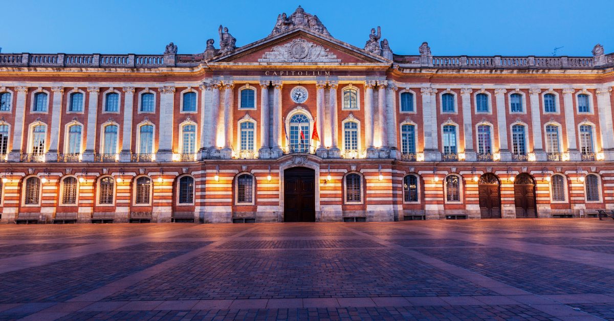 Un grand bâtiment historique de Toulouse avec une façade symétrique illuminée au crépuscule. La structure présente des rangées de fenêtres cintrées, des détails architecturaux complexes et une horloge centrale au-dessus de l'entrée. Le premier plan présente une place spacieuse et pavée rappelant un salon élégant.