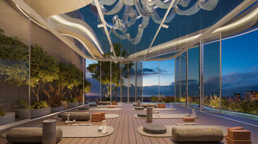 The luxurious rooftop yoga studio at Allura Residences features an open glass ceiling and walls, revealing panoramic views of the city at dusk. The space is adorned with trees, plants, yoga mats and cushions carefully arranged on a wooden deck, creating a serene, modern atmosphere.