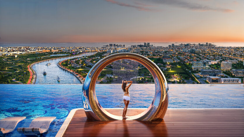 Une personne se tient debout à l'intérieur d'une structure circulaire au bord d'une piscine à débordement, surplombant un paysage urbain tentaculaire et une rivière pendant le coucher du soleil. Des chaises longues sont partiellement immergées dans la piscine à gauche. Le ciel est orné des teintes chaudes du soleil couchant.