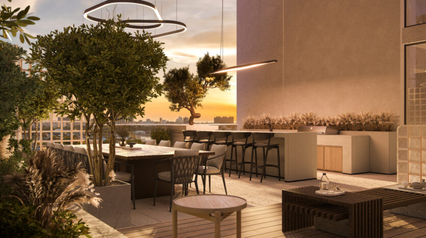 At sunset, the Allura Residences rooftop terrace is cozy and features a dining area with chairs and table, surrounded by lush greenery. Modern lighting fixtures hang overhead and a stylish bar with stools sits in the background. The warm colors of the setting sun enhance the welcoming ambience of these luxury apartments.