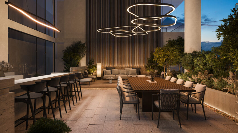 Allura Residences offers a modern outdoor dining area with a long wooden table and upholstered chairs, next to a bar counter with stools. Elegant circular light fixtures hang above, and the space is surrounded by greenery, plants and vertical wall decorations. The atmosphere is serene and elegant.