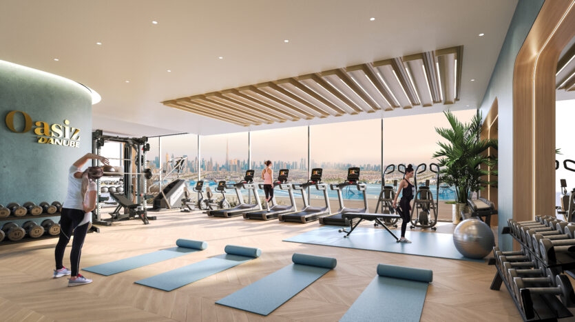 Une salle de sport moderne avec de grandes fenêtres offrant une vue sur le paysage urbain. L'équipement comprend des tapis de course, des vélos stationnaires, des poids et des tapis de yoga. Une personne utilise une bande de résistance au premier plan tandis que trois personnes utilisent les tapis de course. « Oasiz Danube » est affiché bien en évidence sur le mur.