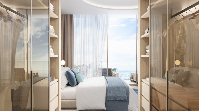 Chambre moderne avec portes de placard en verre reflétant la pièce. Un grand lit avec des draps bleus et blancs fait face à des baies vitrées. Des rideaux transparents révèlent légèrement le balcon du Beach Walk 3, offrant une vue panoramique sur l'océan et une chaise, le tout sous un ciel lumineux.