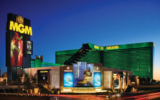 Vue extérieure en soirée de l'hôtel et casino MGM Grand de Las Vegas, illuminé de lumières vertes et dorées. Des panneaux d'affichage et des enseignes bien visibles affichent diverses publicités, notamment un clin d'œil à leurs activités à Abu Dhabi. Une grande statue de lion doré se dresse majestueusement à l'entrée principale.