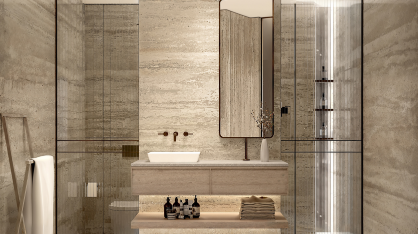 Une salle de bains moderne au One Beverly Dubai avec des murs et un sol en marbre beige. Elle dispose d'un miroir rectangulaire au-dessus d'une coiffeuse en bois clair avec un lavabo blanc. Sous la coiffeuse se trouvent des serviettes pliées et des produits de toilette. À droite se trouve une cabine de douche en verre, tandis qu'à gauche se trouve une échelle porte-serviettes.