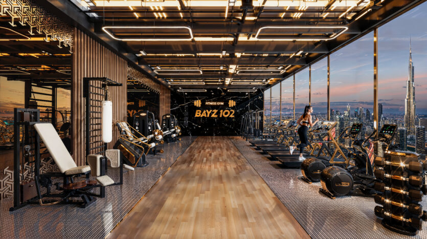 Une salle de sport moderne au Bayz 102 avec parquet et murs en verre offre une vue imprenable sur l'horizon. Elle dispose de divers équipements d'exercice, notamment des tapis de course, des vélos stationnaires et des appareils de musculation. Une personne sur un tapis de course profite du paysage pittoresque.