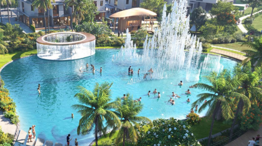 Une piscine extérieure avec une grande fontaine au centre, entourée d'une végétation luxuriante et de palmiers. Les gens nagent et jouent dans l'eau. À proximité, une structure architecturale moderne et ronde fournit de l'ombre. La scène animée regorge d'activité, incarnant l'esprit vivant de Sun City.