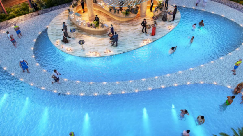 Vue aérienne d'une piscine aux eaux bleues illuminées, entourée de personnes nageant et se relaxant. La pièce maîtresse de la piscine est une terrasse en bois ornée de guirlandes lumineuses et d'un coin bar. Des allées de verdure et de pierre encadrent la piscine, renforçant l'atmosphère sereine de Sun City.