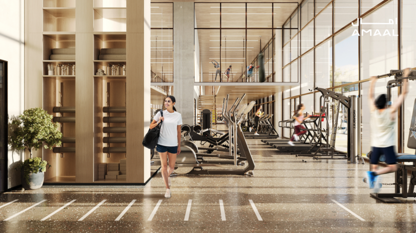 Dans la salle de sport moderne AMAAL 8, les gens s'entraînent sur diverses machines. Une femme traverse l'espace en tenant un sac. La salle est dotée de grandes fenêtres, d'accents en bois et d'un éclairage naturel éclatant. Une plante en pot décore une étagère près de l'entrée, ajoutant une touche de verdure à ce cadre dynamique.