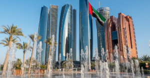 L&#039;immagine mostra i moderni grattacieli con facciate in vetro riflettente di Abu Dhabi, sullo sfondo di un cielo azzurro e limpido. In primo piano, fontane d&#039;acqua e palme arricchiscono il panorama, mentre la bandiera degli Emirati Arabi Uniti sventola in primo piano al centro, catturando l&#039;essenza di questa città iconica in un contesto accessibile.