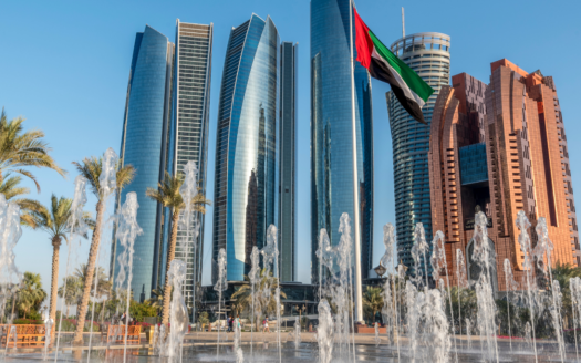 L'image montre des gratte-ciels modernes aux façades en verre réfléchissant à Abu Dhabi, sur fond de ciel bleu clair. Au premier plan, des fontaines à eau et des palmiers enrichissent la vue, tandis que le drapeau des Émirats arabes unis flotte bien en évidence au centre, capturant l'essence de cette ville emblématique dans un cadre abordable.