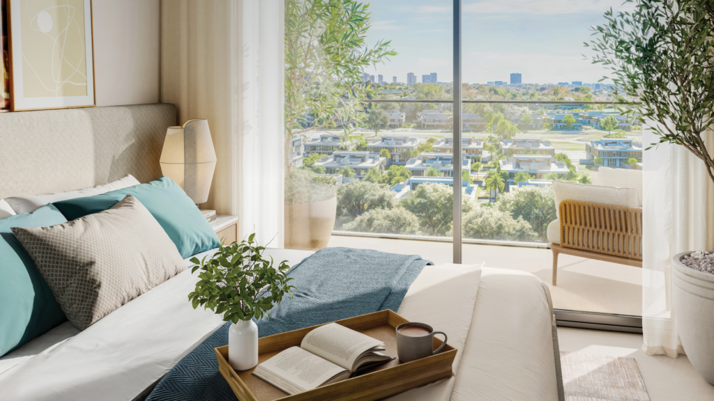 Une chambre ensoleillée de la résidence Vida comprend un lit soigneusement fait avec des oreillers bleu sarcelle et beige. Un plateau avec un livre ouvert et une tasse à café repose de manière accueillante sur le lit. De grandes portes vitrées révèlent un paysage serein et verdoyant et un paysage urbain lointain, tandis qu'une plante en pot ajoute du charme dans le coin.