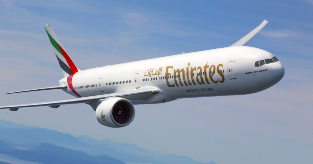 Un Boeing 777 d'Emirates, une splendide création de l'aviation moderne, s'élève dans un ciel bleu clair. L'avion est doté d'une carrosserie blanche ornée du logo Emirates, tandis que ses moteurs bourdonnent sous les ailes. Des montagnes enneigées s'étendent au loin.