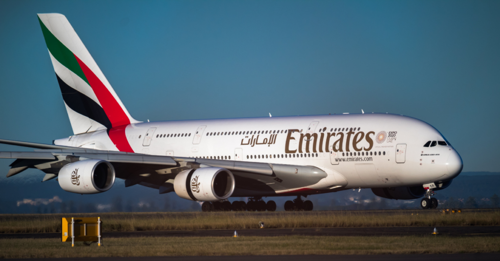 Un Airbus A380 d'Emirates, une création de la célèbre compagnie aérienne, roule sur une piste. L'avion brille de blanc, orné du logo Emirates et d'une livrée mettant en valeur les couleurs du drapeau des Émirats arabes unis sur sa queue. Un ciel bleu clair et un paysage lointain offrent une toile de fond époustouflante.